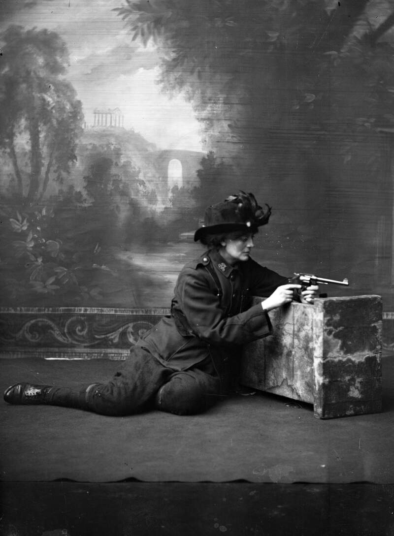 [Countess Constance Markievicz in uniform, kneeling against a studio prop holding a gun, studio full-length portrait]