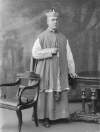 [Most Rev. William Mac Neely standing between table and chair, full-length portrait]