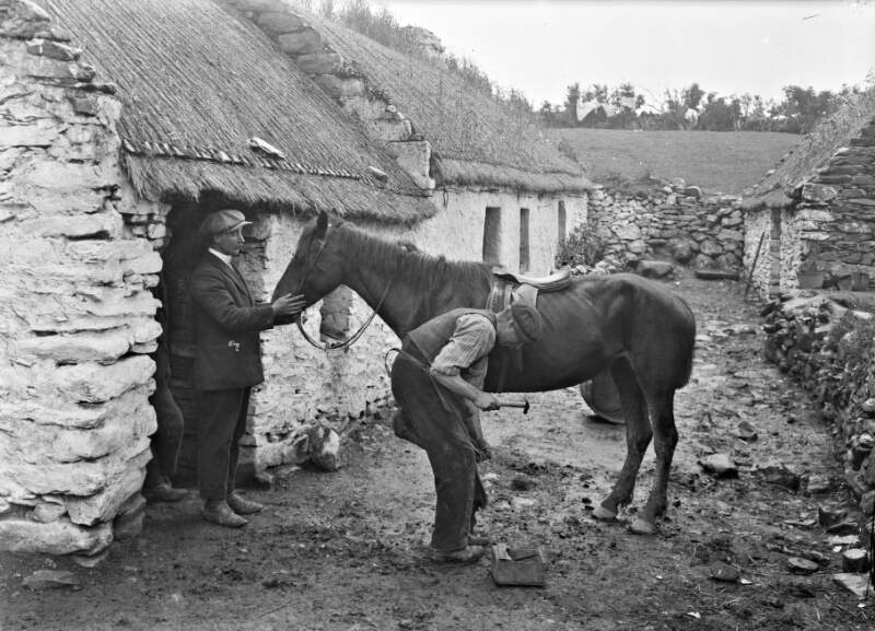 [Blacksmith shoeing horse outside thatched cottages, Ireland]