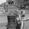 [Young girl reading newspaper beside ornate ironwork bollards, Parnell Monument, O'Connell Street, Dublin]