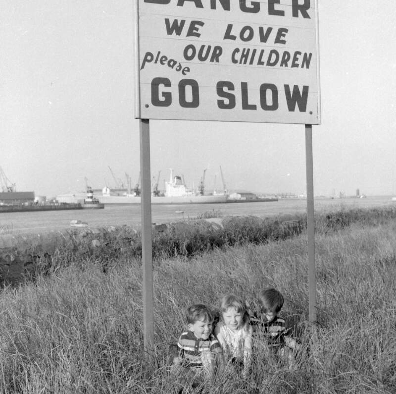 [Children seated under traffic sign on shore, ships in background, Pigeon House Road, Ringsend, Dublin]