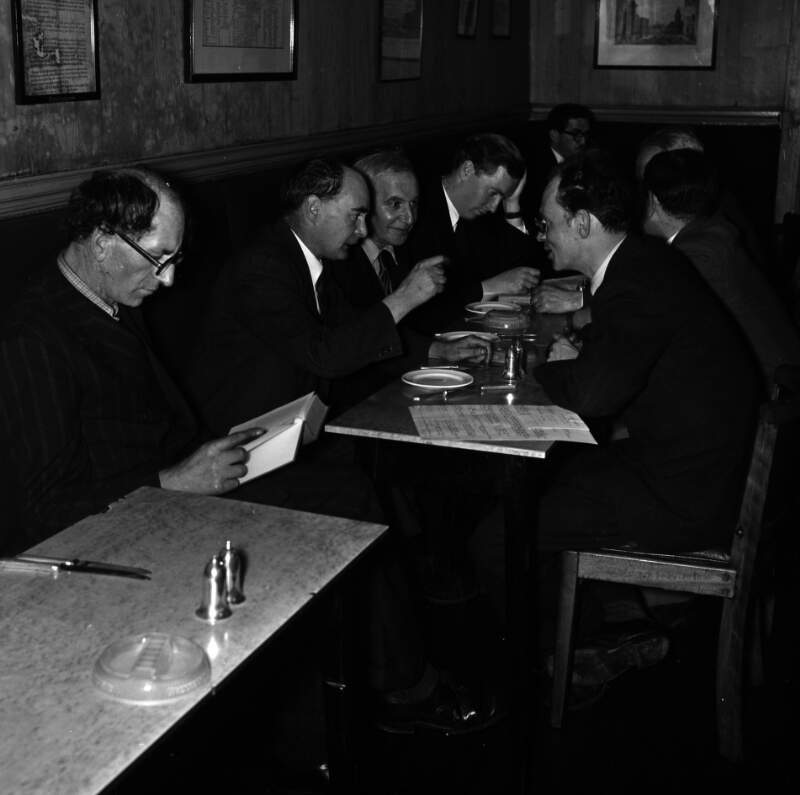 [Patrick Kavanagh reading book, Flann O'Brien gesturing across table, Con Leventhal, John Ryan with head in hands, unidentified man in background, Anthony Cronin at Davy Byrnes pub, Bloomsday, Duke Street, Dublin]