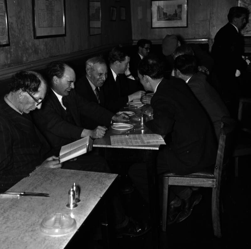 [Patrick Kavanagh reading book, Flann O'Brien laughing, Con Leventhal, John Ryan with head in hands, unidentified man in background, others facing away from camera, Davy Byrnes pub, Bloomsday, Duke Street, Dublin]