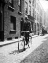 [Man wearing hat, cycling bike on cobbled street]