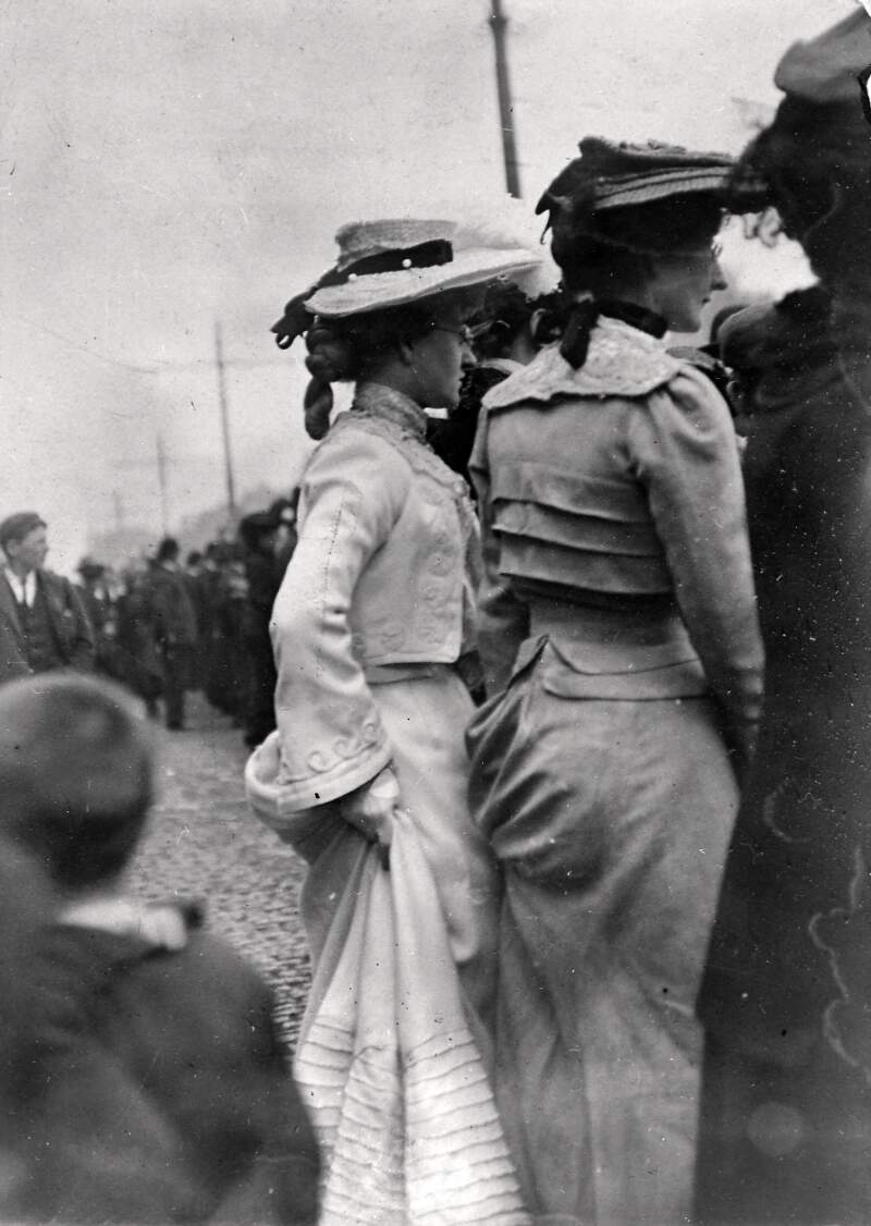 [Two women standing in a crowd on a street, with their backs to the camera]