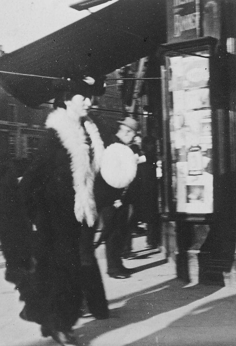 [Woman with white fur collar and muff, walking past stationery shop]