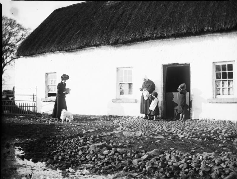 [Edith Dillon carrying a box camera, approaching woman and two children standing outside thatched cottage]