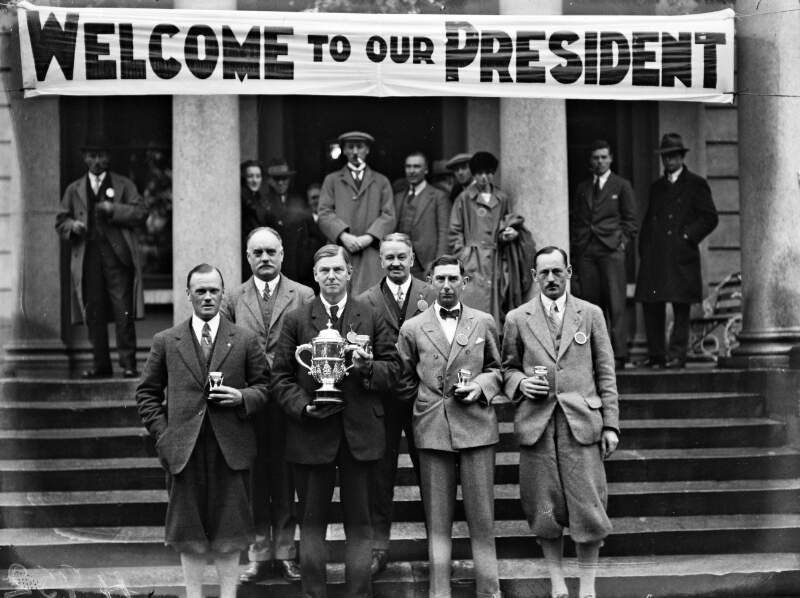 [Rotary convention, Killarney. Group with trophy and cups on the steps of the Great Southern Hotel. Banner reads "Welcome to our president"].