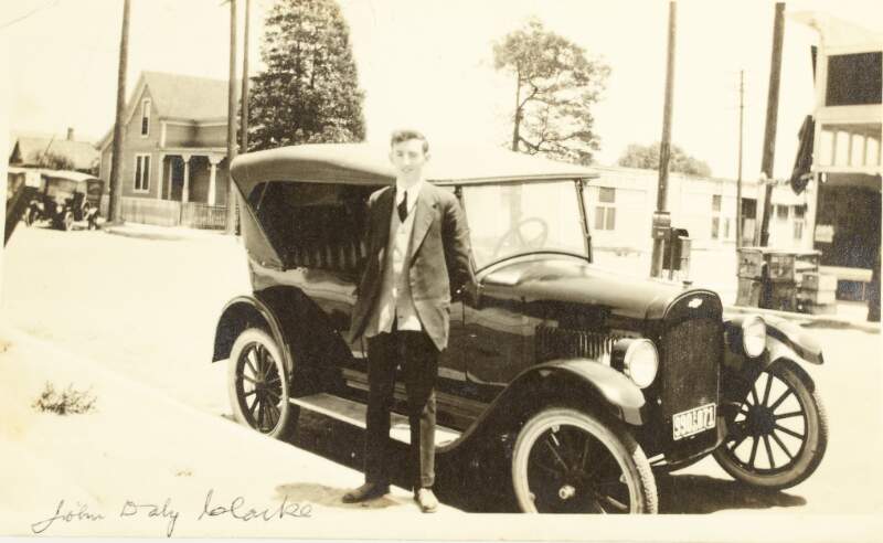 [John Daly Clarke, full length portrait of him standing alongside a motor car]