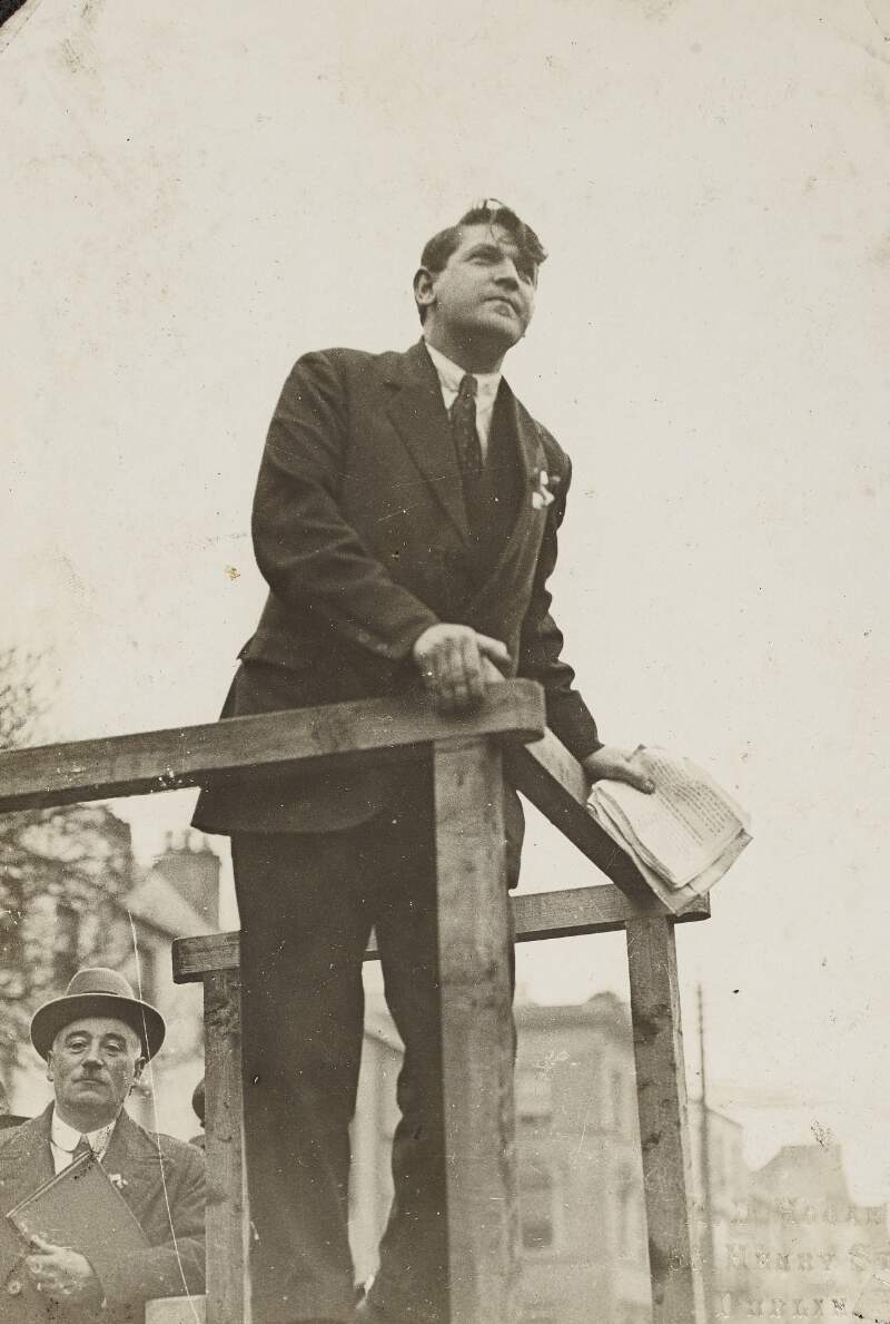 [View of Michael Collins speaking from podium : with an unidentified person in the background]