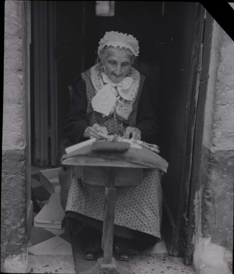 Old woman in doorway, working needles/crochet.