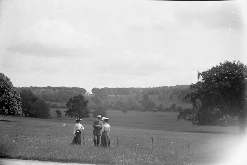 [Two women and a man standing by fence - parkland in distance. Note large gateway in background.]