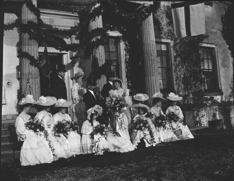 [Bridal party on steps of Clonbrock House. Portico decorated with flowers, women holding large bouquets of flowers and wearing wide brimmed hats, Augusta Dillon standing on steps.]