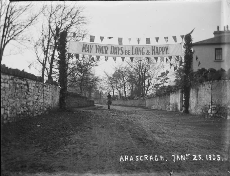 [Ahascragh, Jan 25, 1905] Banner across road in village of Ahascragh - it reads "May your days be long and happy". Man on horseback in distance.]