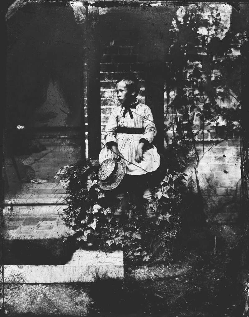 [Young boy in pinafore, seated on ledge of photograph house, holding straw hat in hand (possibly Robert Edward Dillon, b. 1869).]
