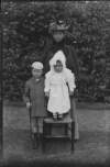 [Young boy, toddler wearing bonnet and embroidery anglaise cloak standing on chair, woman stands behind them, she wears check skirt with shawl-like coat and hat with mantilla.]