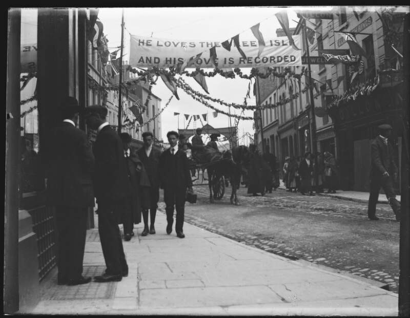 [Street decorated with bunting and banners. Banner reads "He loves the green isle and his love is recorded". Crowds walking on pavements, similar to Clonbrock 409 (Galway city on occasion of Edward VII's visit).]