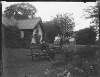 [A.C.C on wheelbarrow seat. Elderly Augusta Dillon on seat in front of photograph house and small glasshouse.]