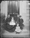 [Family group on doorstep, includes woman with four children, two toddlers wearing tunics.]