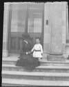 [Woman and little boy on steps of large house. He wears dress or tunic. Woman wears ruffled dress with straw bonnet tilted forwards.]