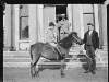 [Little boy on pony in front of Clonbrock House. Man holding reins (possibly an employee) and other one on porch smoking pipe. Boy wears a pinstriped blazer.]