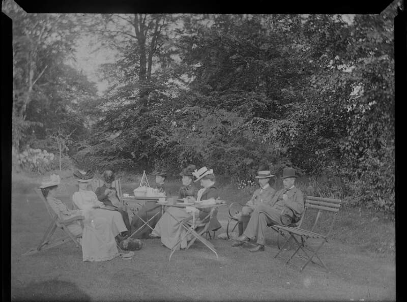 [Group (six men and two women) having tea on lawn, tables set with china, women wearing large hats decorated with bows.]