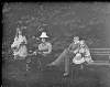 [Group (six men and two women) having tea on lawn, tables set with china, women wearing large hats decorated with bows.]