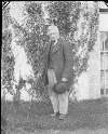 [Portrait of elderly man with walking stick outside ivy-covered house.]