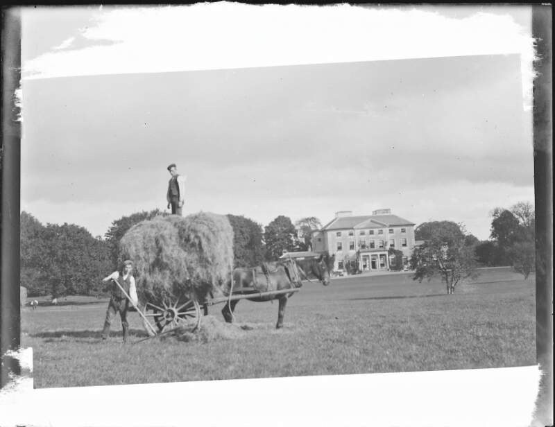 [Hay-making on Clonbrock Demesne, shows horse with cart full of hay. One man standing on cart, another raking hay. House visible in the distance.]