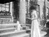 [Bobs - Sept. Dog standing on hind legs on porch of Clonbrock House, Edith Dillon standing in front of him holding saucer of water. Window boxes in background. Edith wears featured hat and plume around her neck.]
