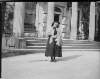 [Bessy Delaney. Elderly woman standing in front of Clonbrock house. She wears a knitted shawl and bonnet. Walking stick on ground . Dishevelled shoes.]