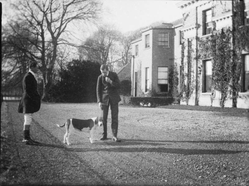 [Two men, one wearing riding outfit, standing on gravel drive in front of house. Beagle dog between them, brick house in background covered in ivy, not Clonbrock House, possibly Oldtown, Naas Co.kildare. Home of De Burgh family,see 816, 818.]