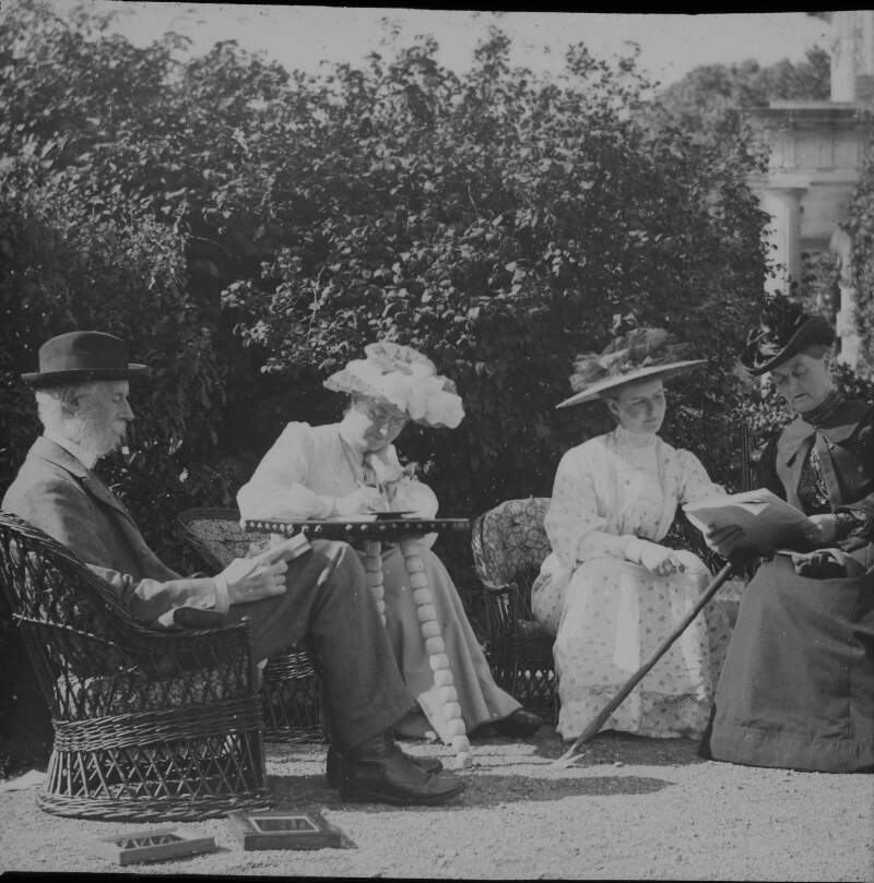 [Group with uncle Ned, 1904. Group seated outdoors on wicker chairs, Clonbrock House in background. Included left to right are Ned (Edward Crofton) smoking a pipe, Georgina Dillon, Ethel or Edith Dillon and Augusta Crofton Dillon. Plate holders on ground.]