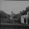 [Ahascragh, Chapel and Twibills, 1903. View of muddy village street, showing thatched cottages, cart and a tree, Church in background. "Twibills" public house or shop in foreground. Local woman visible in distance.]