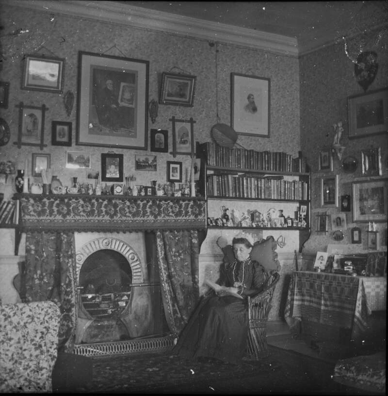 [Mamas Room. Interior of small cluttered room, woman sitting by fireside, row of books, wall of portraits and photographs, ornaments on mantelpiece.]