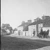 [Ahascragh and waggonette 1903. Ahascragh main street, showing thatched cottages and shops. Wagonette in foreground, driver wearing livery and top hat. Woman and child visible outside shop.]