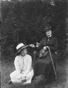 [Augusta Crofton Dillon seated on chair, holds gardening gloves in her hands, also carries walking stick. Woman seated on ground nearby, foliage shrubs in background.]