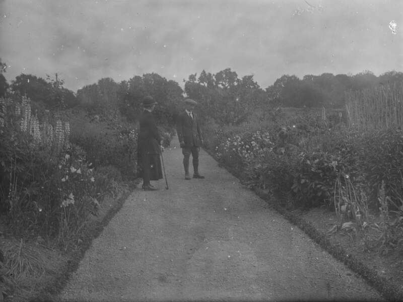 [Elderly man and woman walking on pathway, flowers on either side of walkway, trees in the distance, Clonbrock.]