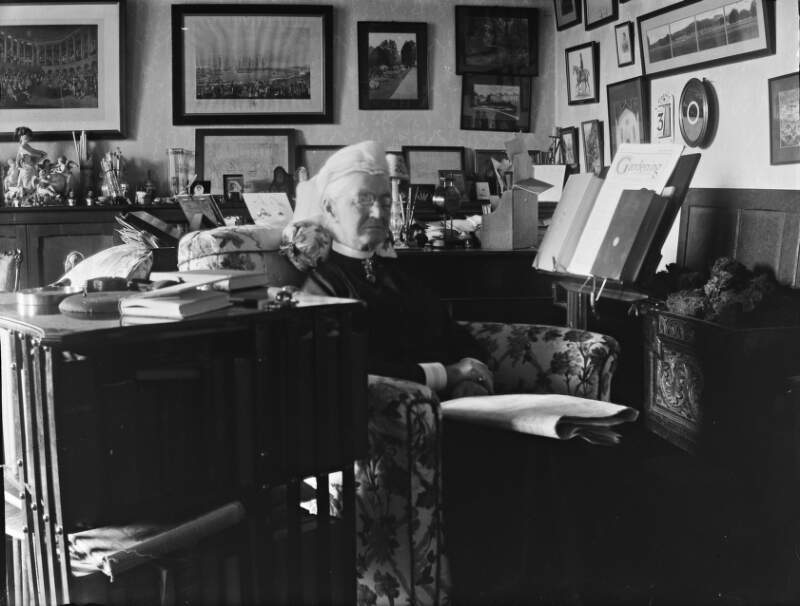 [Interior of cluttered room, Augusta Crofton Dillon seated on armchair, surrounded by ornaments. Many paintings and photographs on wall, also shows reading stand with garden magazine and two books.]