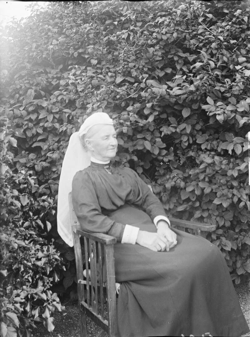 [Augusta Crofton Dillon seated outdoors, Large bush/shrub in background, Augusta wears a long scarf style head dress, similar image to Clonbrock 1168.]