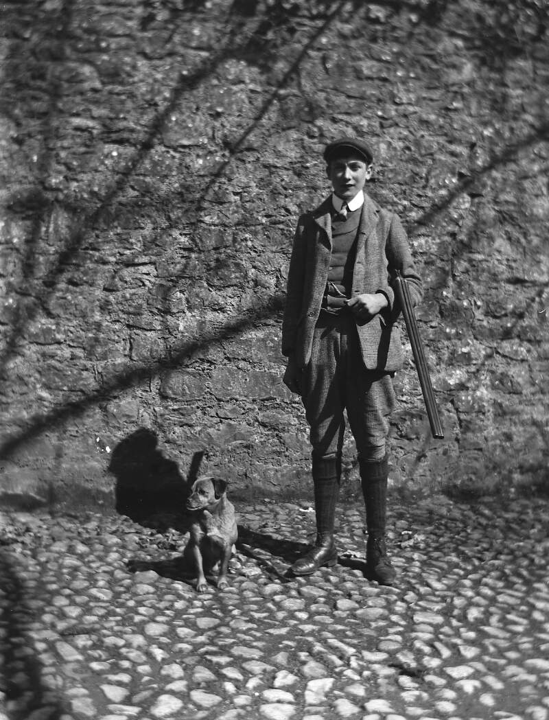 [Young man wearing hunting style outfit standing in cobbled yard, small beside hay. Young man carries rifle, similar image Clonbrock 1270.]