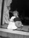 [1907. Toddler sitting on step wears embroidery anglaise pinafore, and holds stuffed teddy bear/doll. Drapes and interior visible in background.]