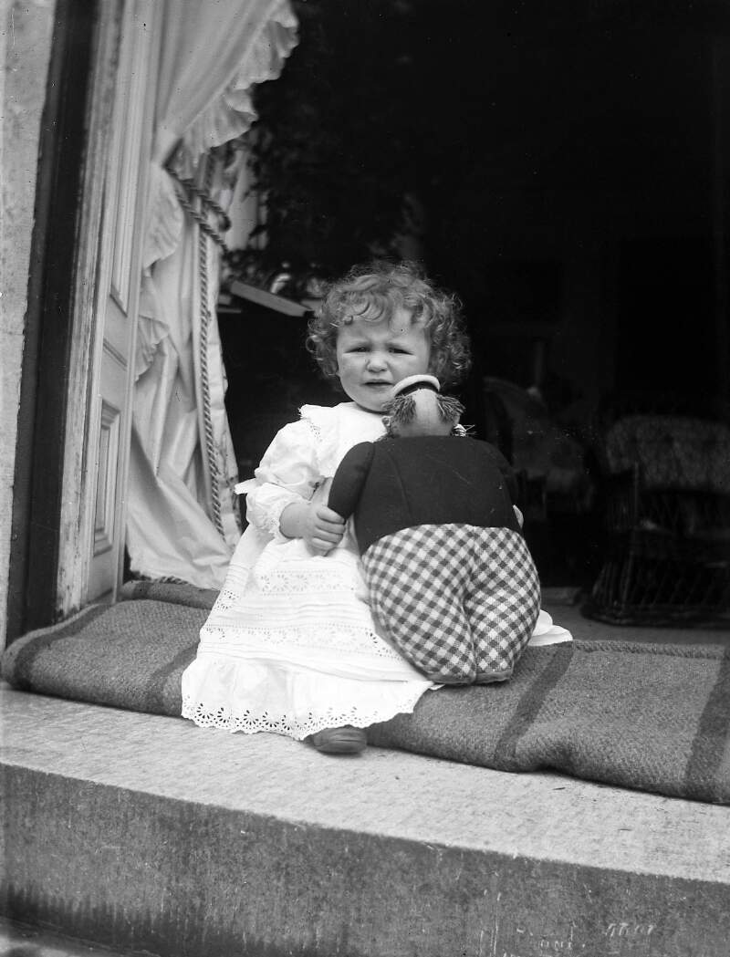 [1907. Toddler sitting on step wears embroidery anglaise pinafore, and holds stuffed teddy bear/doll. Drapes and interior visible in background.]