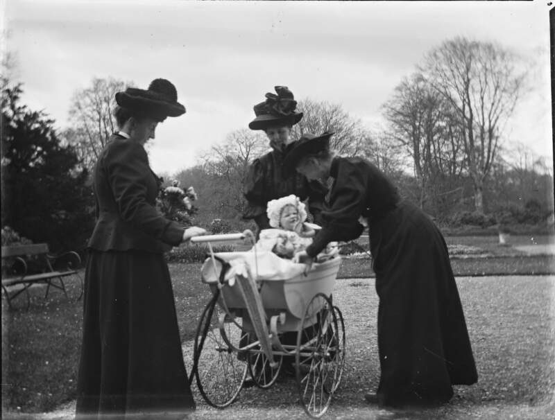 [Two women looking on as Augusta Crofton plays with Ursula Mahon in her pram.]