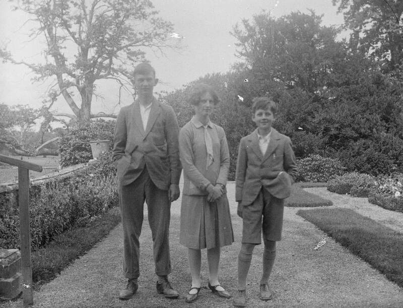 [A couple with a young boy in short pants standing on path with trees and plants in the background.]