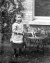 [Young girl standing by wicker table with a basket on top, outside photograph house.]