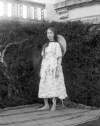 [Young girl standing barefoot on blanket in garden, balcony to house just visible in the background.]