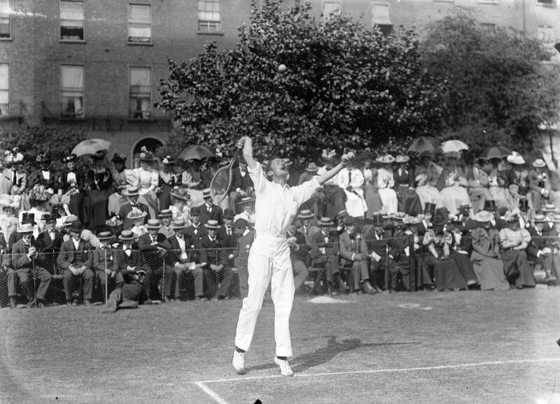 [Tennis player hitting a high ball with arms outstrched, people lining the court in the background.]