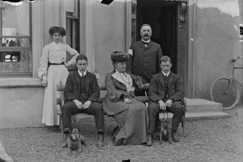 [Group portrait of two women and three men and two dogs in front of house.]