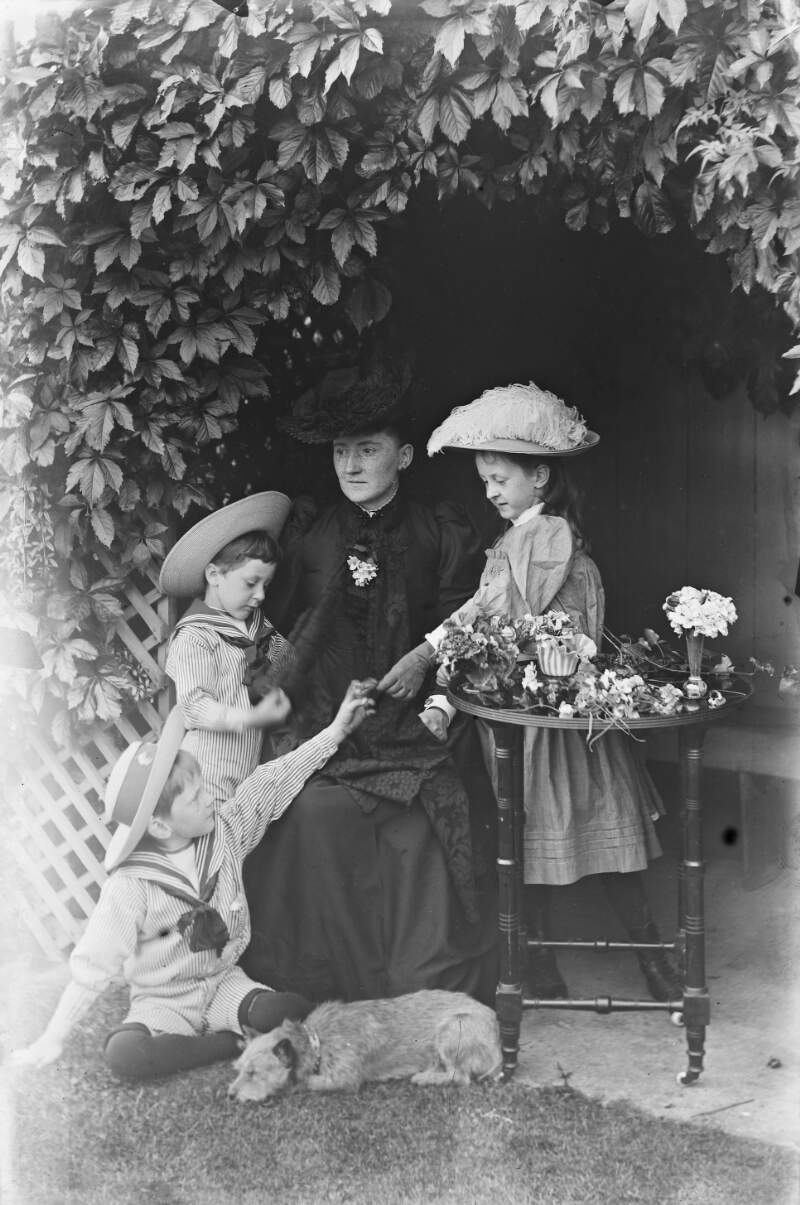 [Woman seated with young girl and two young boys dressed in sailor outfits with hat, with a small table covered with loose flowers, and dog sleeping nearby.]
